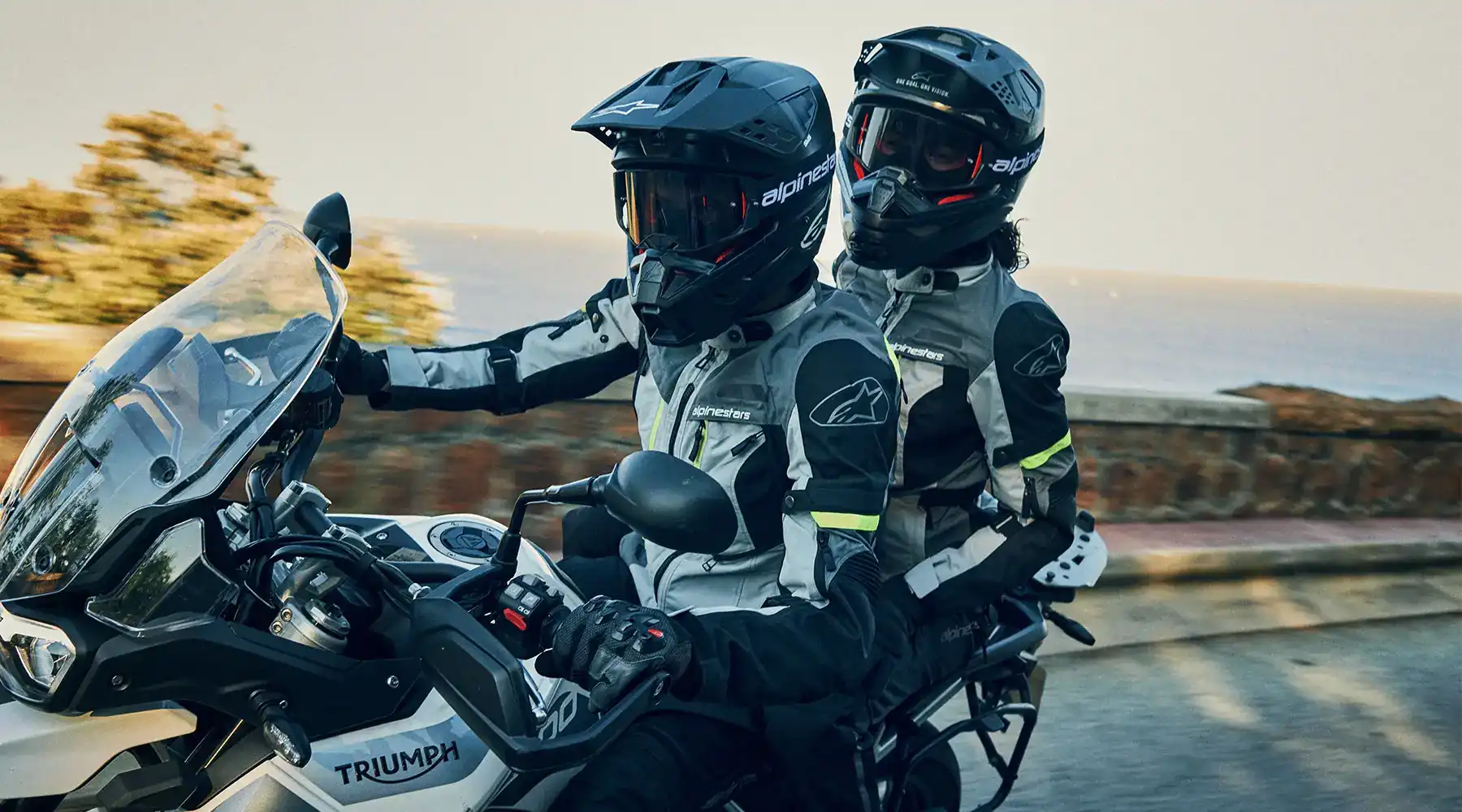 Dos personas con cascos y chaquetas Alpinestars en una motocicleta Triumph, viajando por una carretera costera.