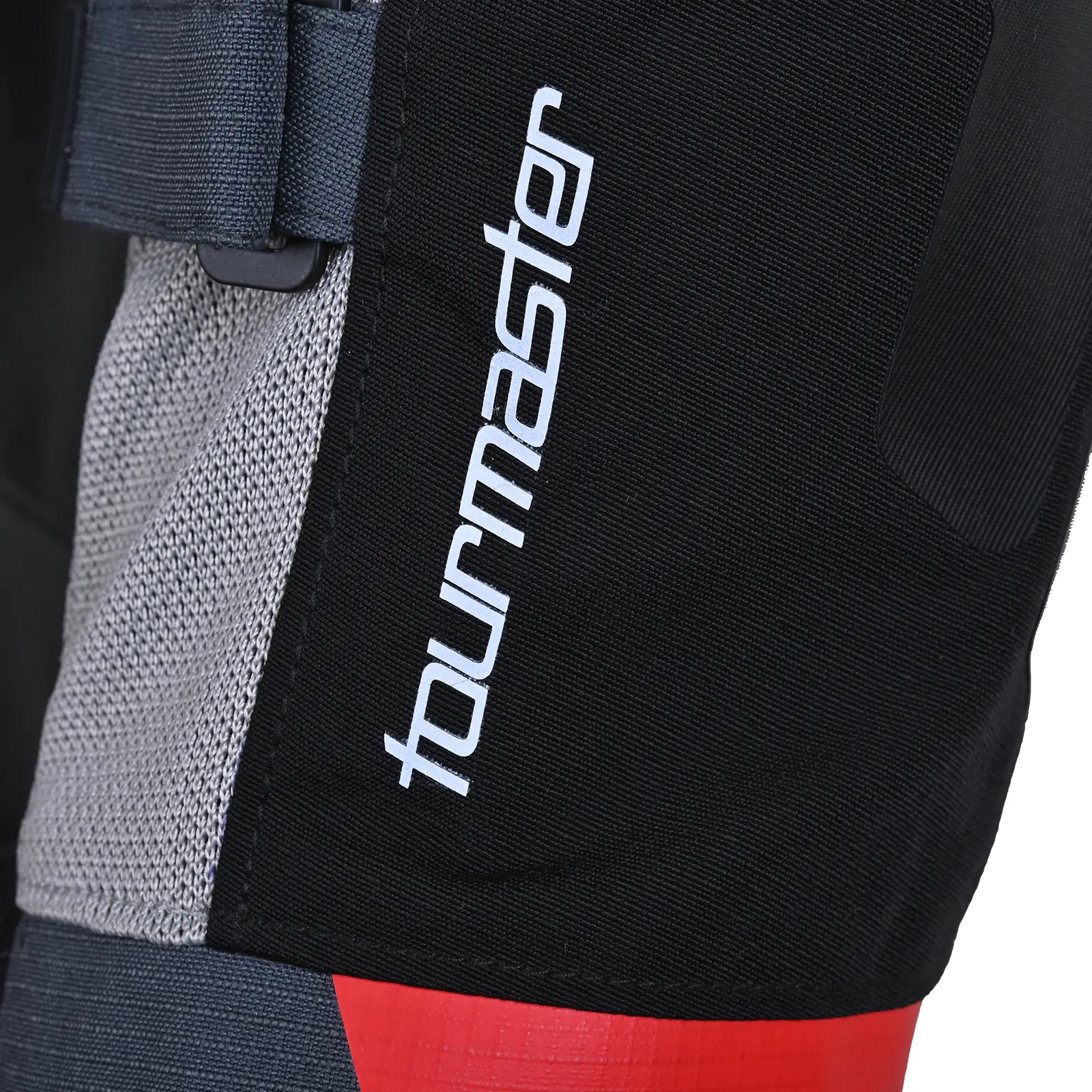 Detalle de la marca Tourmaster en equipamiento de motocicleta, con logo blanco sobre tela negra, inserción de malla gris y ajuste de correa.