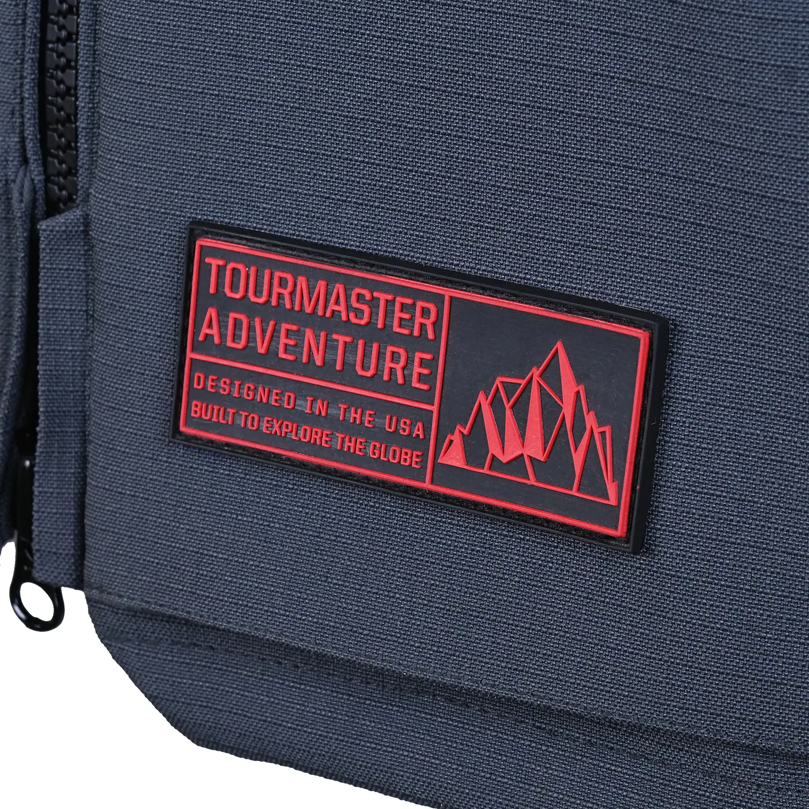 Parche rectangular negro con borde rojo y texto en rojo 'TOURMASTER ADVENTURE' y 'DESIGNED IN THE USA BUILT TO EXPLORE THE GLOBE'. Incluye gráfico de montañas rojas. Cosido sobre tela gris oscuro texturizada.