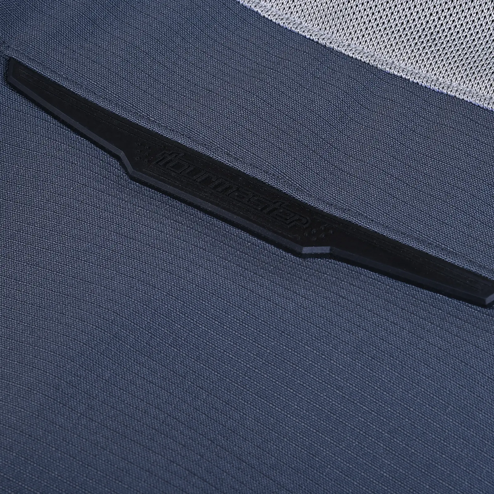 Primer plano del logo Tourmaster en relieve sobre tejido textil de color azul oscuro, con textura visible y parte superior de malla gris.