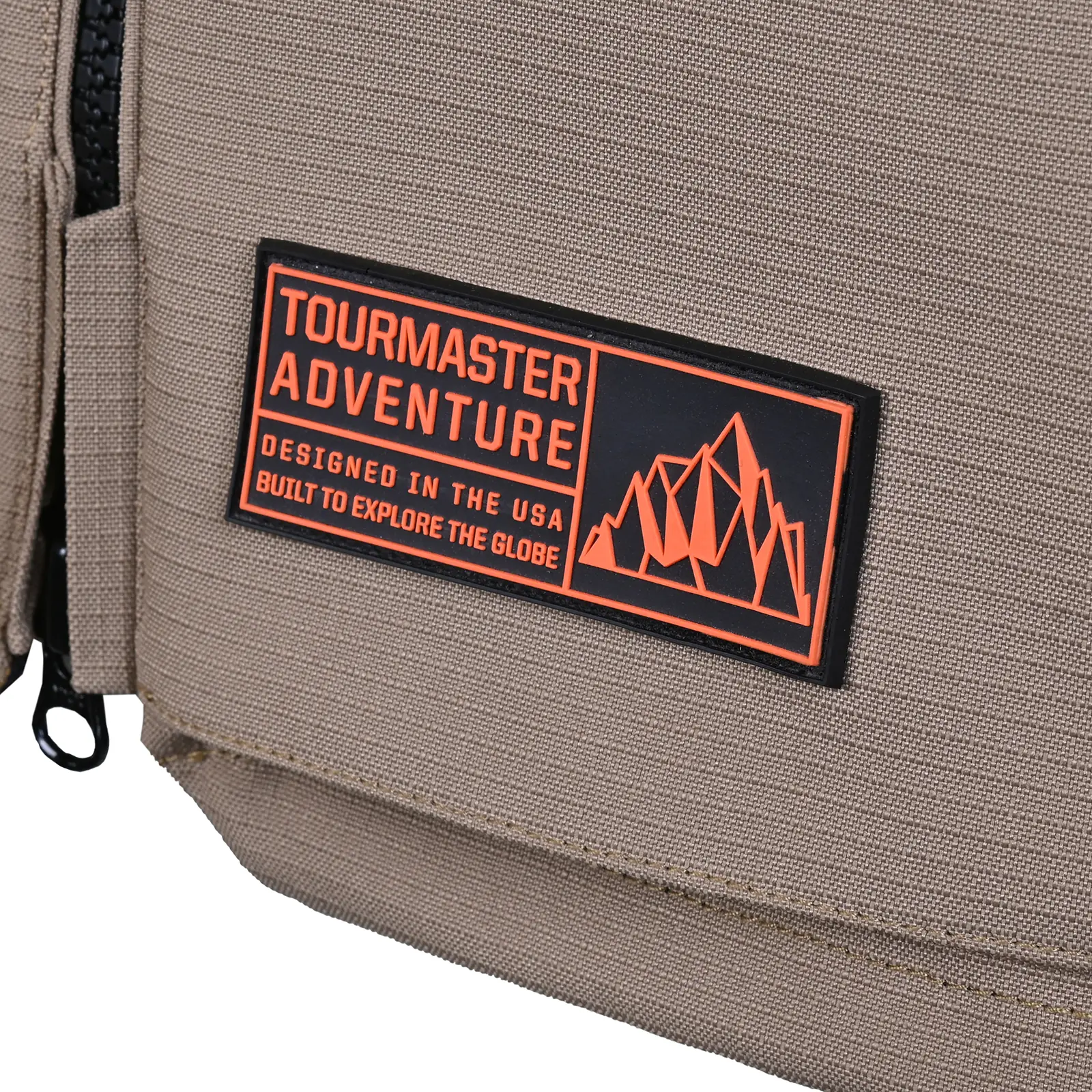 Parche rectangular negro con texto naranja 'TOURMASTER ADVENTURE', 'DESIGNED IN THE USA', 'BUILT TO EXPLORE THE GLOBE' y gráfico de montañas. Cosido sobre tela textil color caqui con cremallera negra visible.