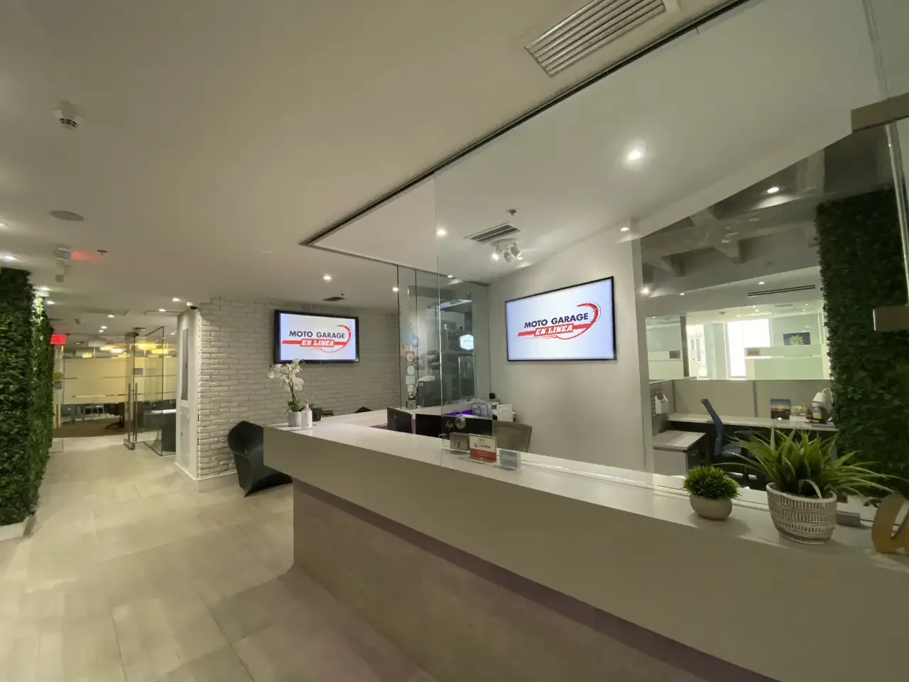 Recepción de oficina moderna con dos pantallas mostrando el logo 'Moto Garage En Línea', paredes de ladrillo blanco, follaje verde artificial y mostrador de atención al cliente.