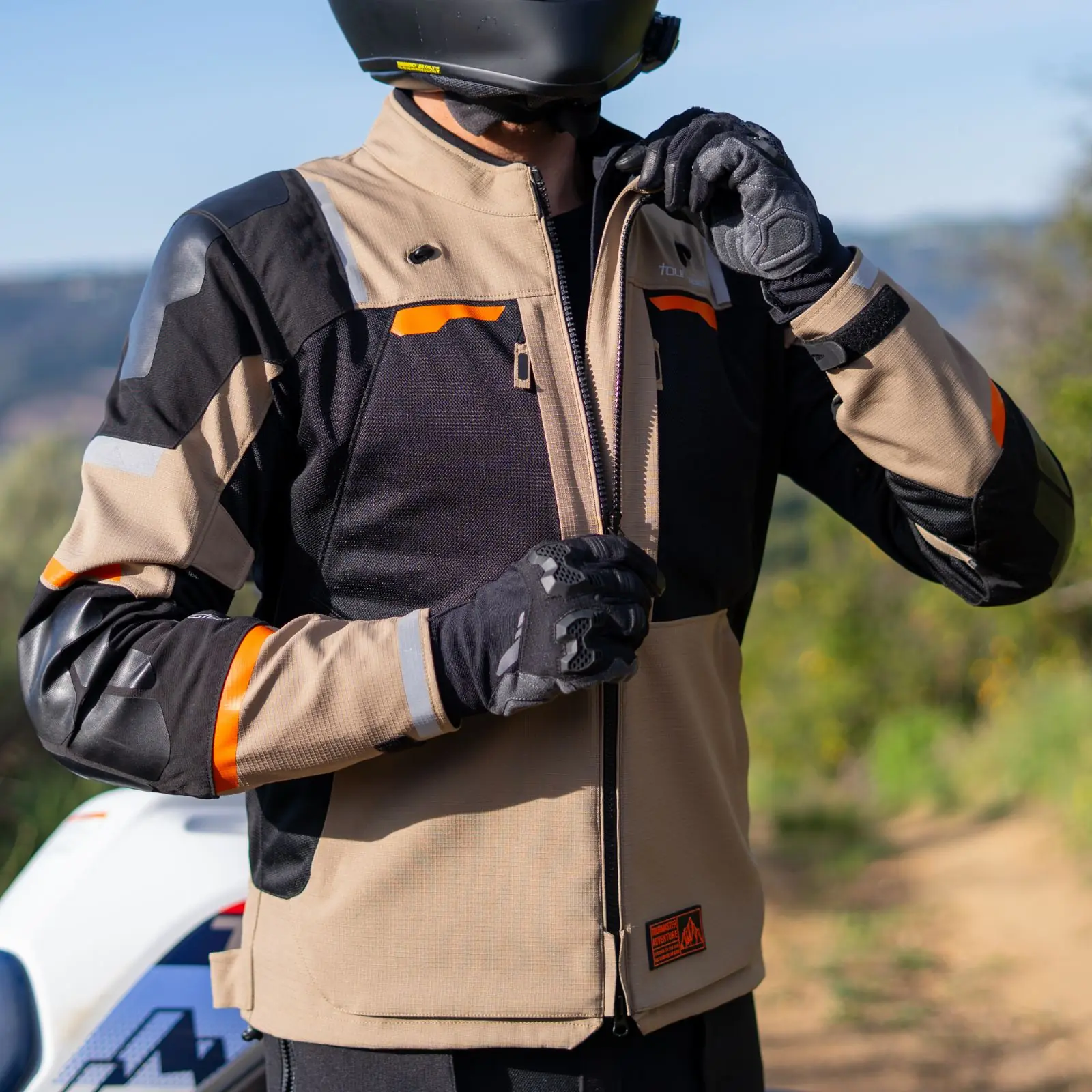 Hombre con casco negro, chaqueta de moto beige, negra y naranja, y guantes, abrochando el cierre frontal de la chaqueta, con una motocicleta blanca visible en el fondo.