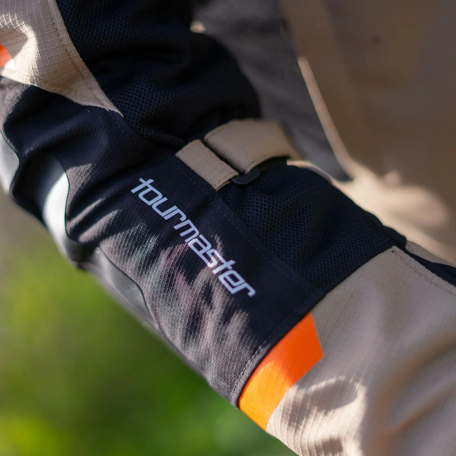 Primer plano de manga de chaqueta Tourmaster con logo blanco, tejidos negro y beige, panel de malla, detalle naranja y correa ajustable.