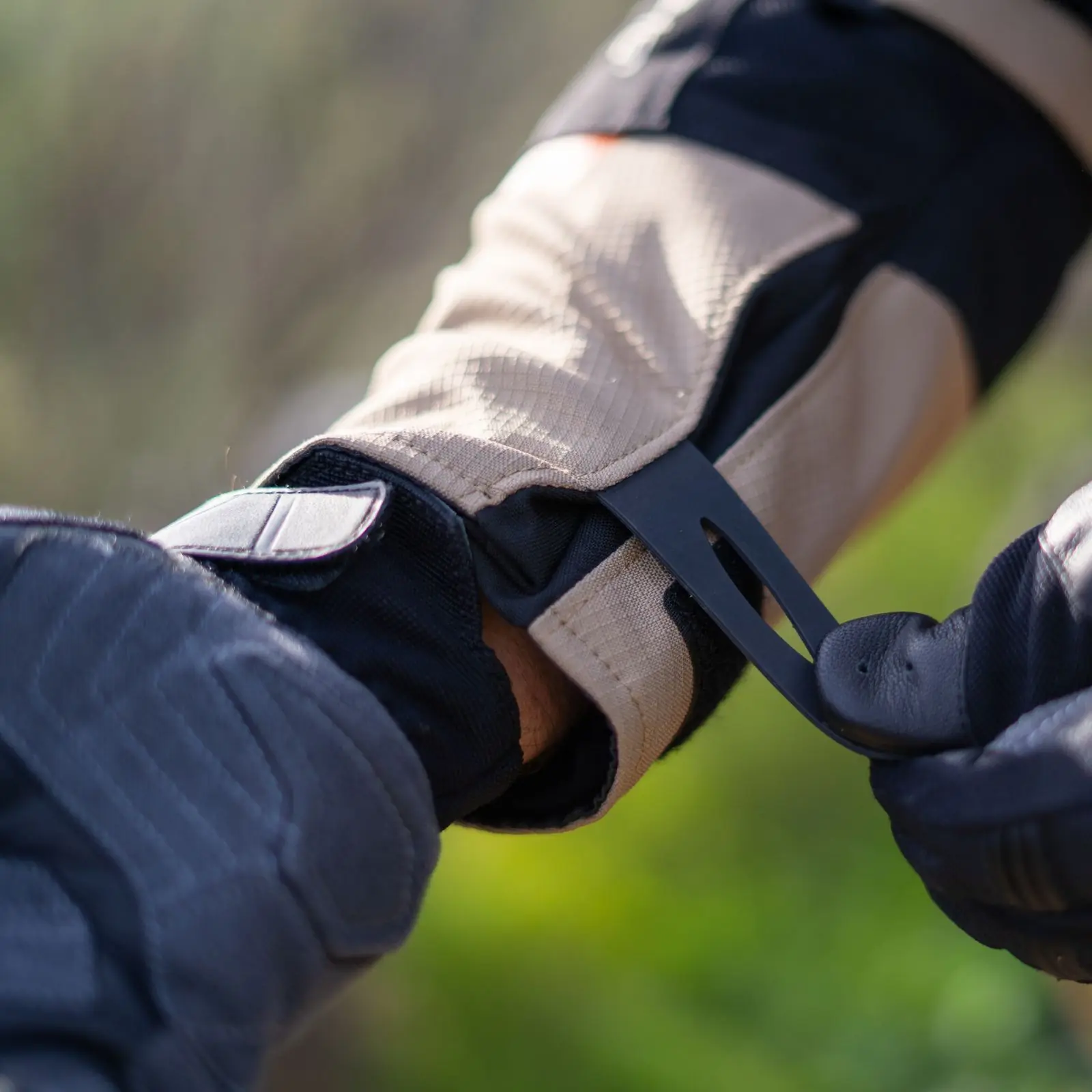 Primer plano de mano con guante de motocicleta gris oscuro ajustando la correa de ajuste de la muñeca de una chaqueta de moto color beige y negro. Fondo verde difuminado.
