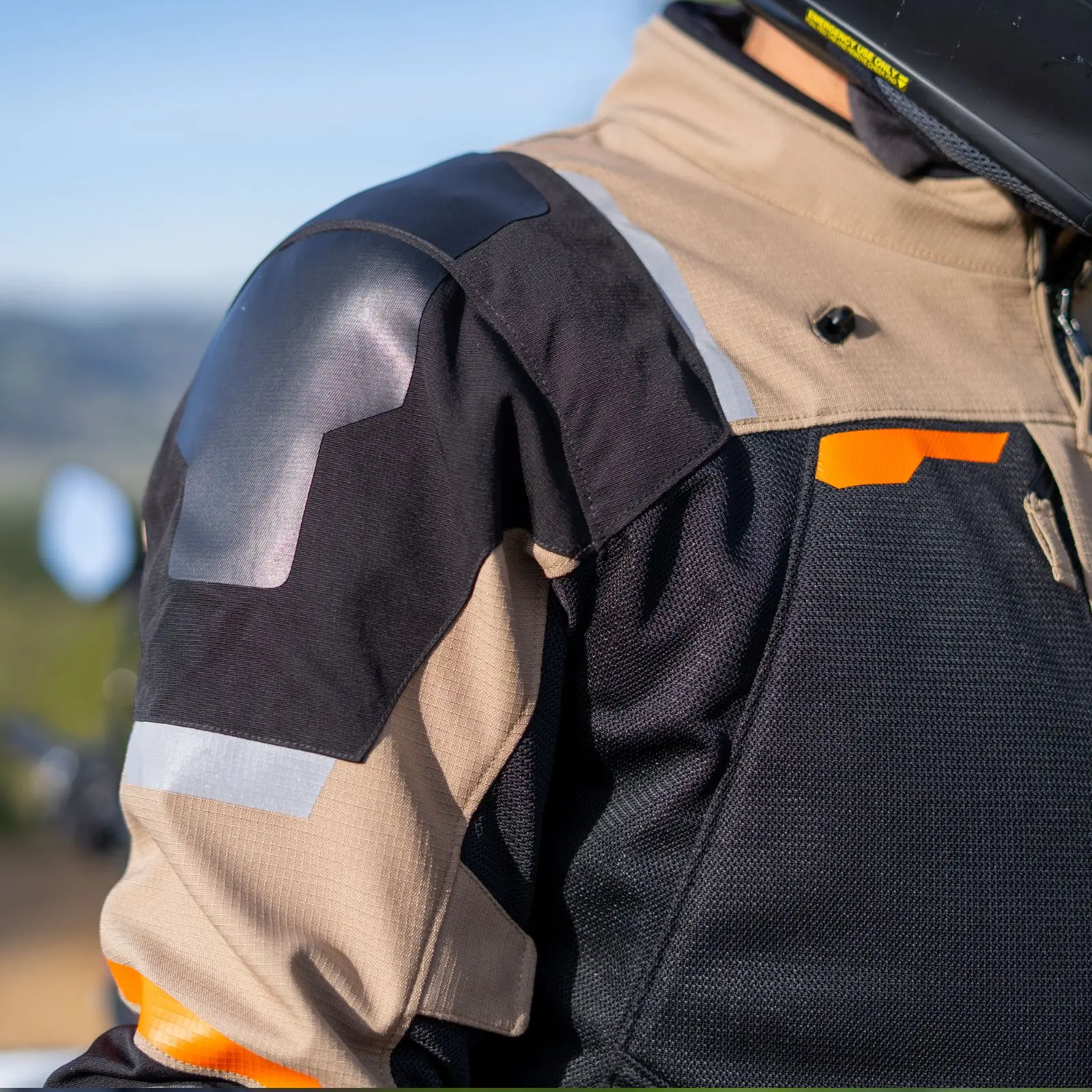 Primer plano de chaqueta de motocicleta en colores beige, negro y naranja. Detalles de tela texturizada, paneles reflectantes y hombrera protectora visible.