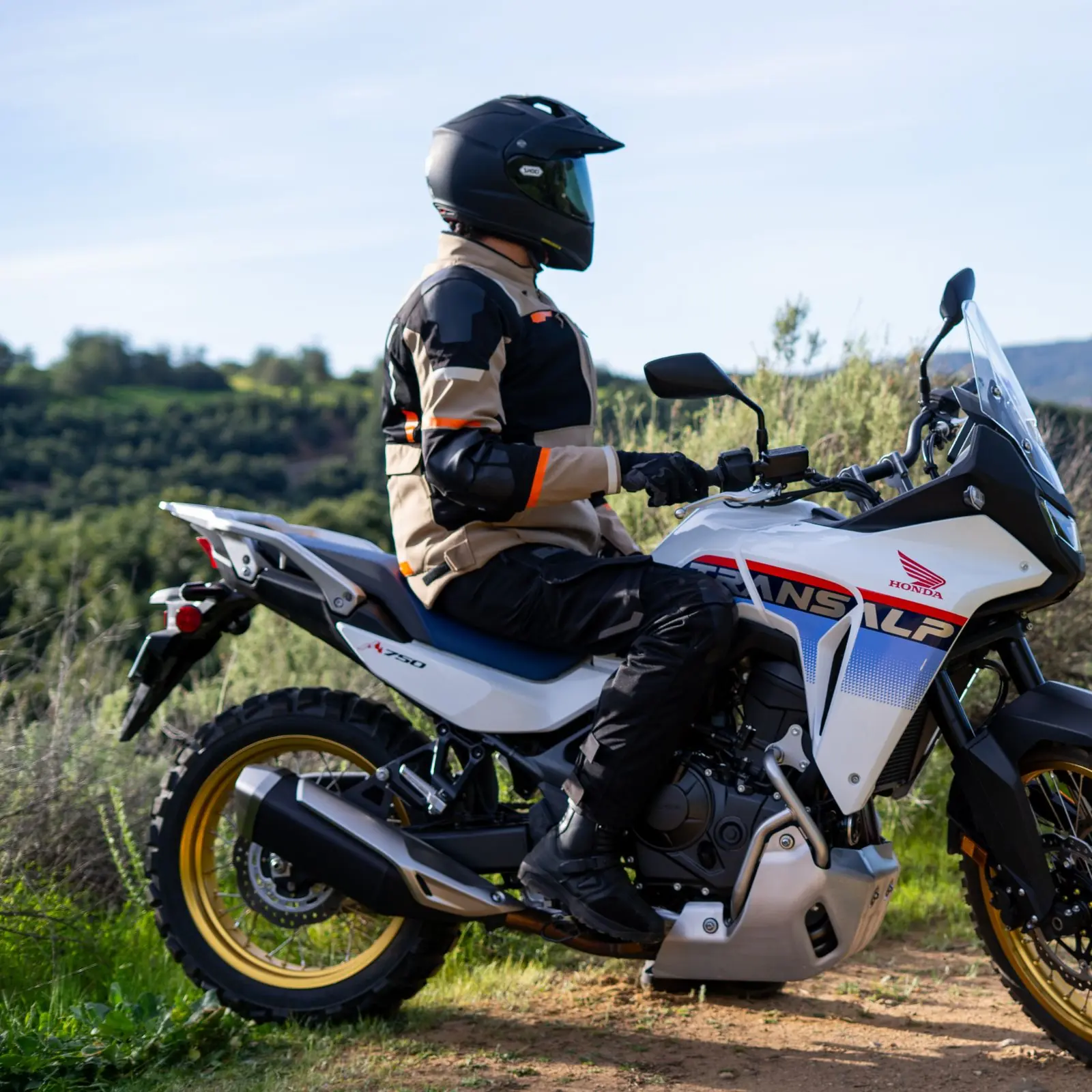 Hombre con casco Shoei negro y equipo de motocross sobre motocicleta Honda Transalp XL750 blanca con llantas doradas en camino de tierra.