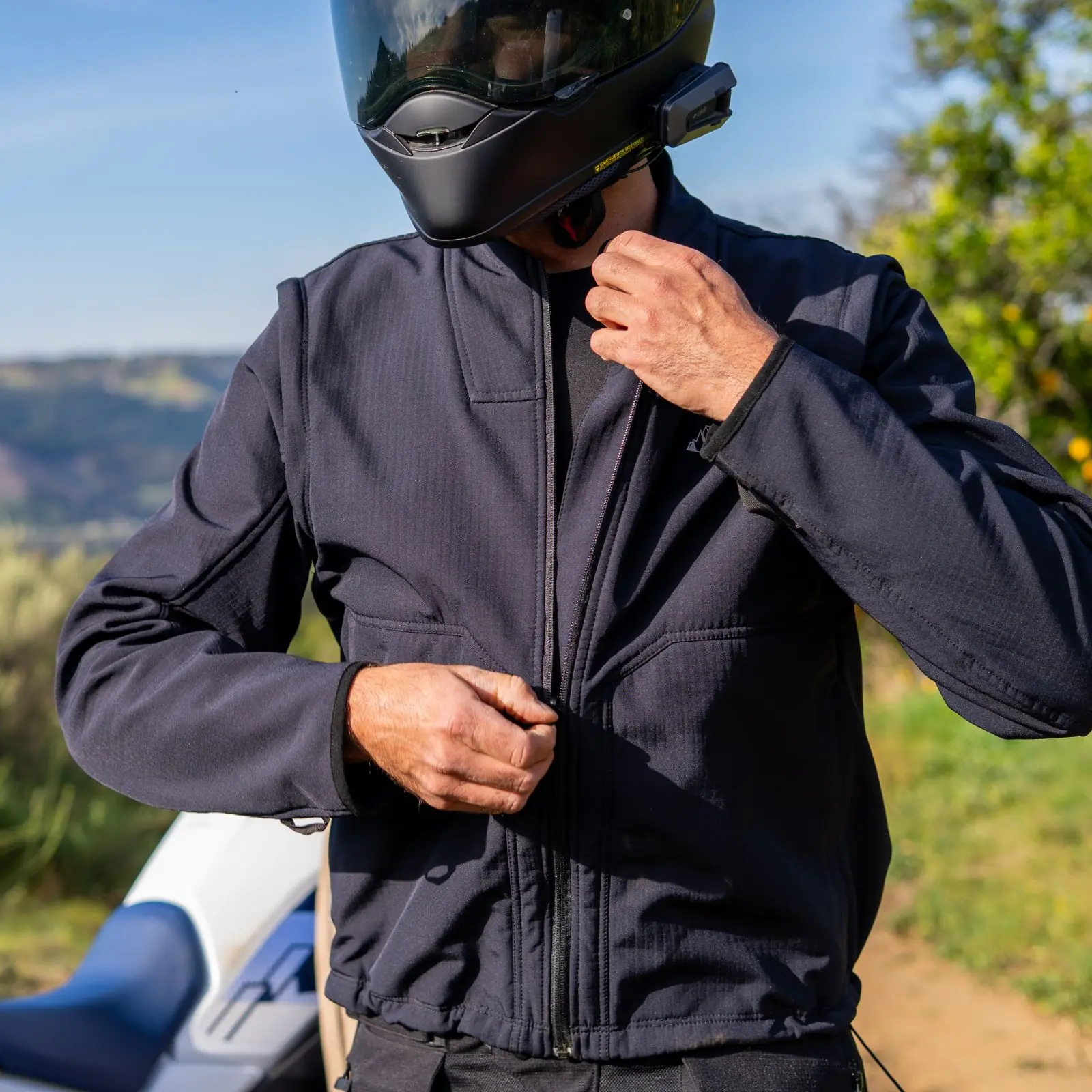 Hombre con casco integral negro y chaqueta de motocicleta oscura abrochando cremallera frontal, en exterior con paisaje montañoso.