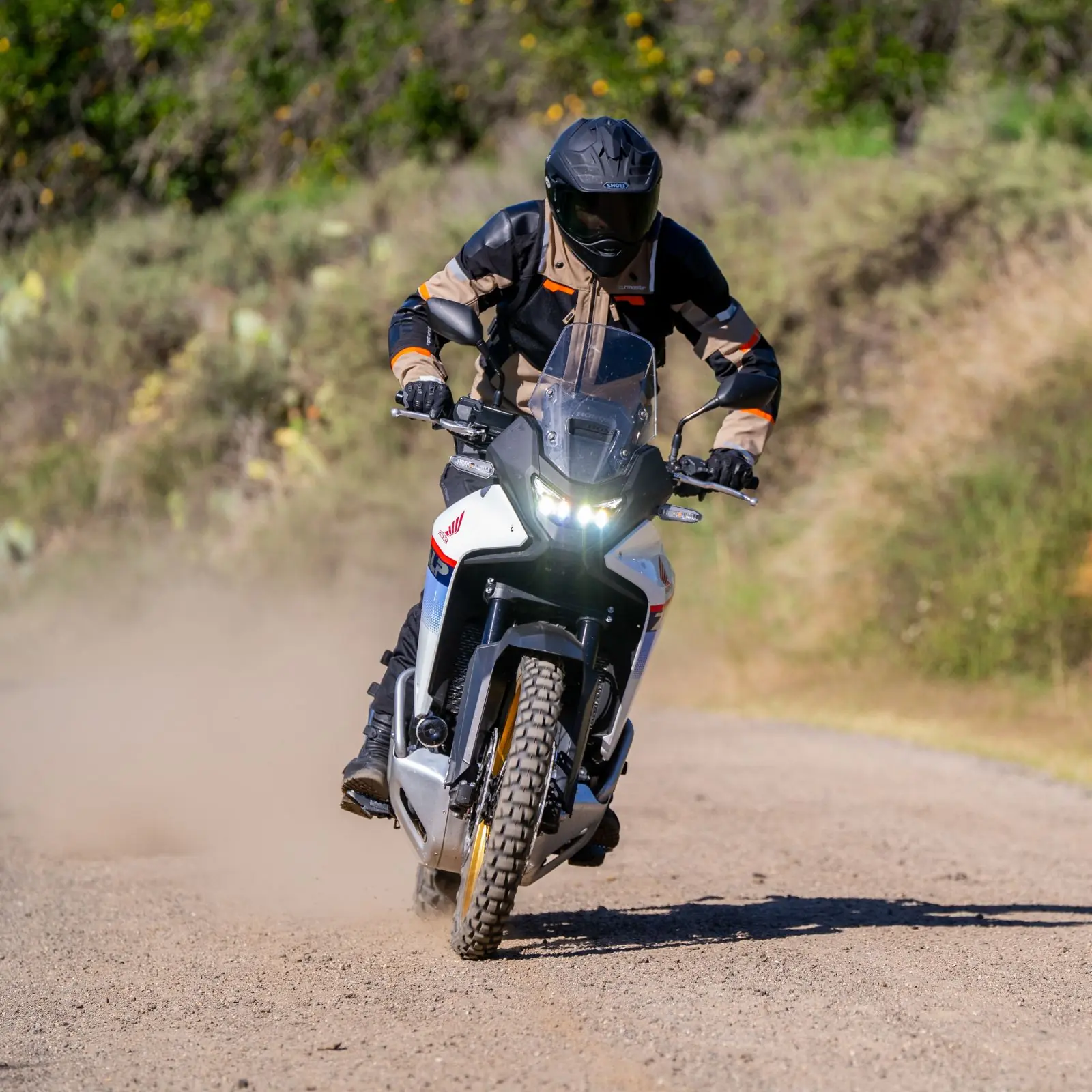 Motociclista con casco negro, chaqueta de aventura beige y negra, guantes, y botas, conduciendo una motocicleta Honda adventure blanca, azul y roja con luces LED encendidas, en un camino de tierra levantando polvo, con vegetación verde borrosa de fondo. Vista frontal-diagonal de la motocicleta en movimiento.