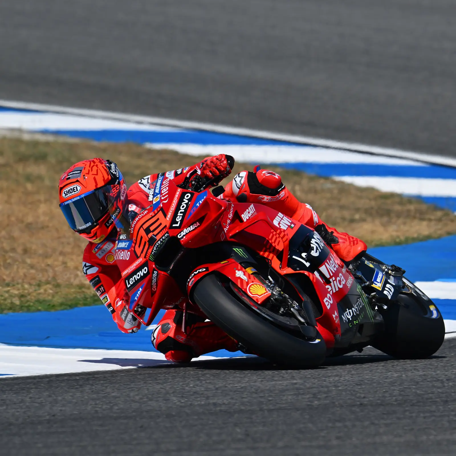 Piloto de MotoGP en motocicleta Ducati Desmosedici GP roja, número 63, inclinándose en una curva de pista. Equipamiento: casco Shoei, traje de carreras rojo con logotipos de patrocinadores (Lenovo, Shell, Monster Energy, Ducati, Michelin). Fondo de circuito de asfalto y cielo azul.