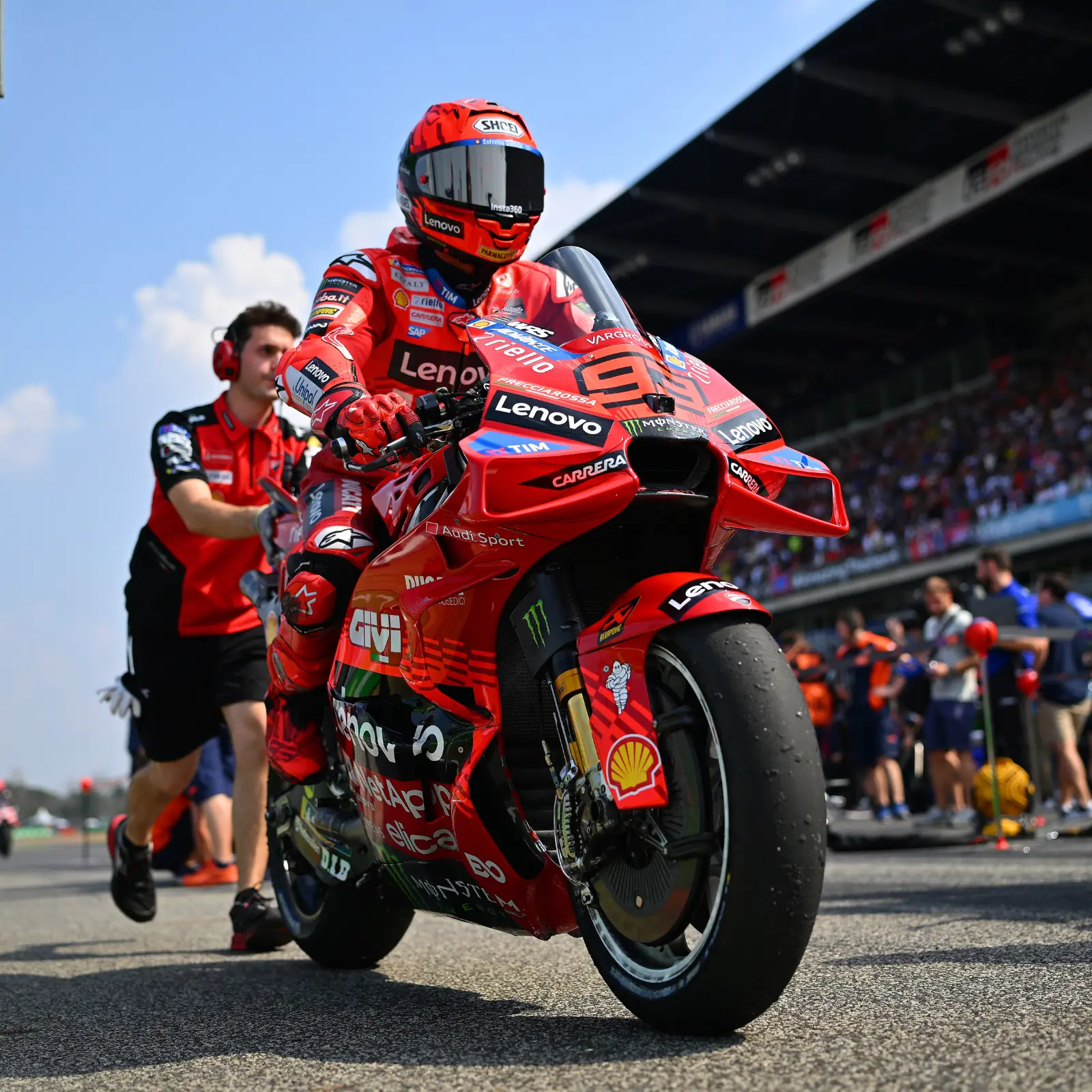 Piloto de MotoGP con traje de carreras rojo y casco Shoei, empujando una motocicleta Ducati roja con múltiples logotipos de patrocinadores en la pista de un circuito de carreras.