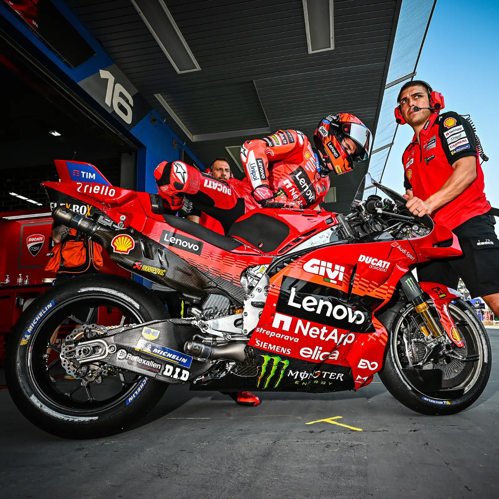 Motocicleta de carreras Ducati Desmosedici GP en paddock con piloto y mecánico, patrocinadores Lenovo, Akrapovic, Michelin, Monster Energy.
