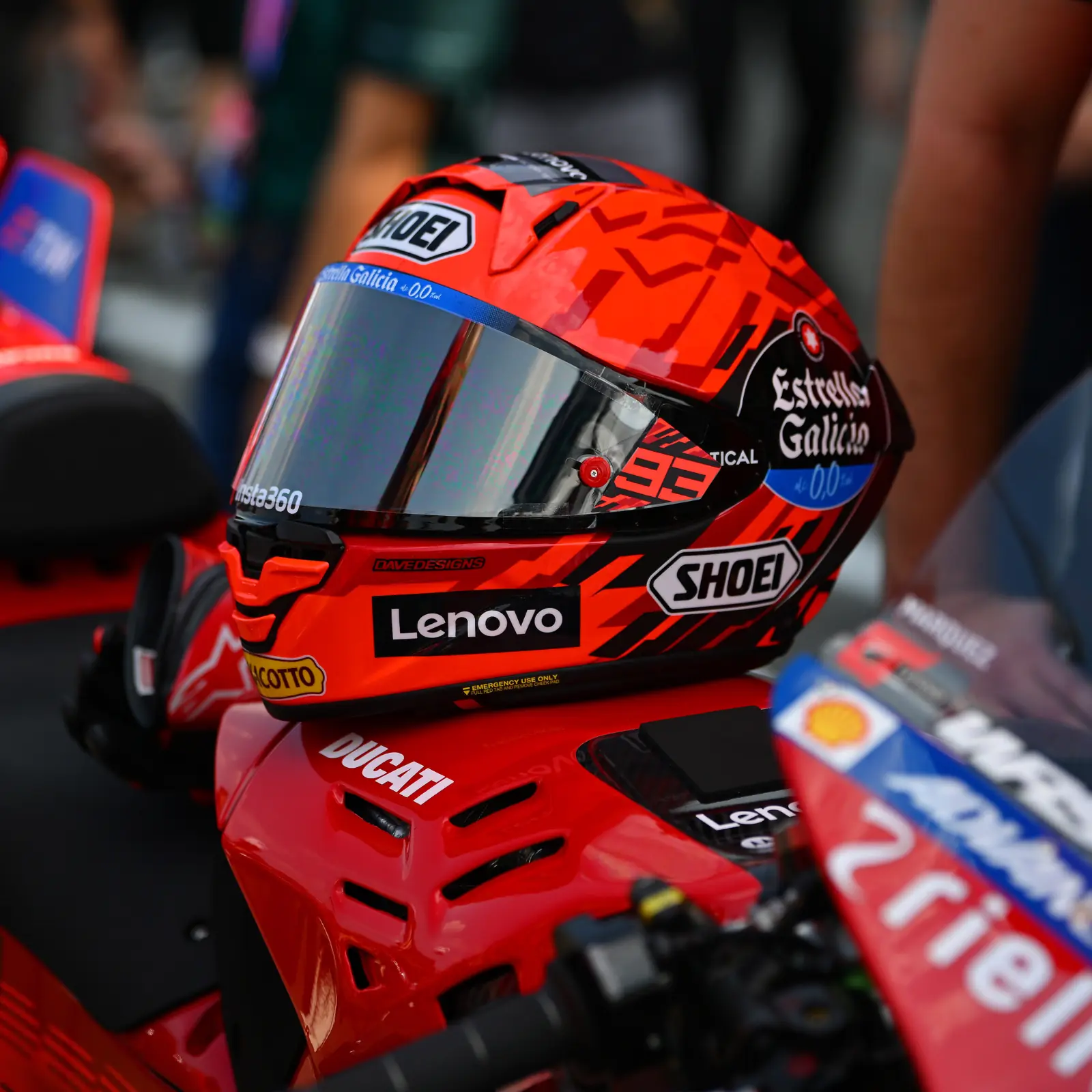 Casco de motocicleta Shoei rojo con logos de patrocinadores (Shoei, Estrellas Galicia 0,0, Lenovo, Insta360, Dave Designs, 93, Vertical, Cotto) apoyado sobre una motocicleta Ducati roja. Visor oscuro.