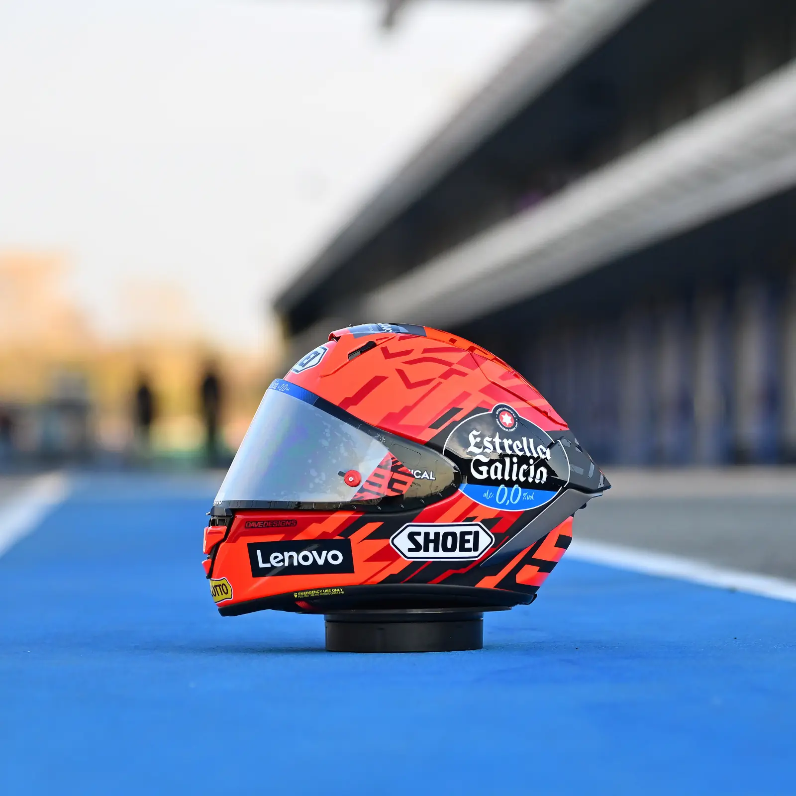 Casco de motocicleta Shoei X-SPR Pro en colores naranja, negro y rojo con patrones geométricos y logotipos de patrocinadores como Lenovo y Estrella Galicia. El casco está sobre una superficie azul que simula una pista de carreras.